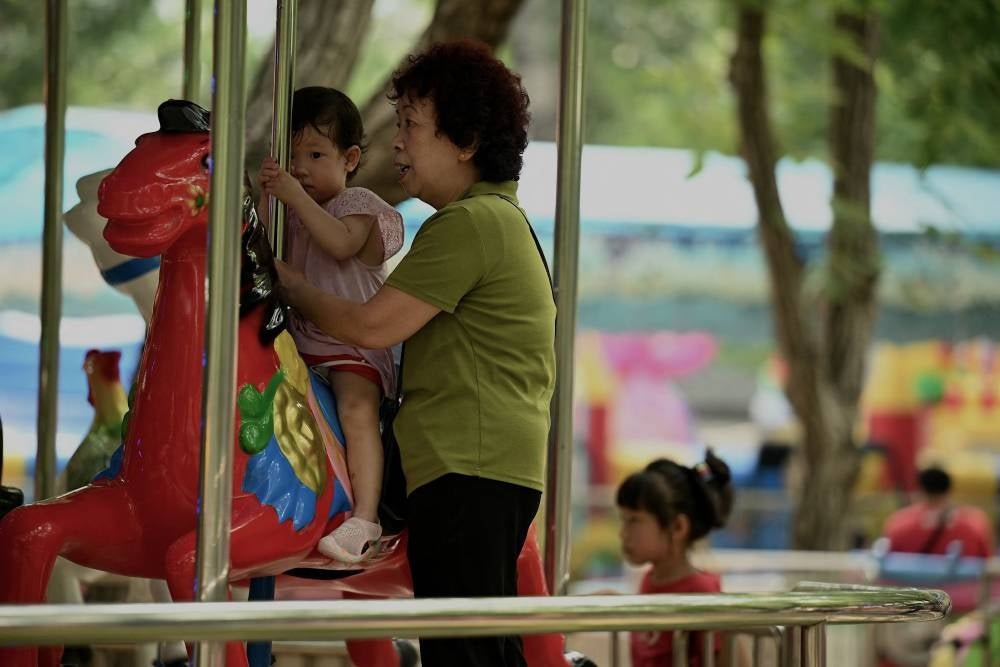 A woman tends to a child riding a merry-go-round at a park in Beijing on August 2, 2022. - China's population will begin to shrink by 2025, officials said, as family sizes grow smaller and citizens age. (Photo by Noel Celis / AFP)