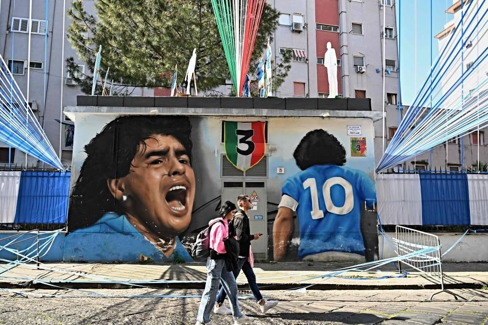 People walk past a graffiti depicting Argentine's football legend Diego Maradona, with flags, banners and a papier mache model of Naples's football players (up), in the colors of Napoli, decorating the Quartieri Case Nuove in central Naples on April 29, on the eve of the Italian Serie A football match between Napoli and Salernitana. - Naples, as the city braces up for its potential first Scudetto championship win in 33 years. An incredible 19 points clear at the top of Serie A, it's surely only a matter of time until southern Italy's biggest club win the Scudetto for the first time since 1990, when Diego Maradona was still strutting his stuff in sight of Mount Vesuvius. - Pic: AFP
