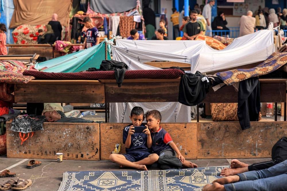 A child sits next to another playing a game on a cell phone in the playground of a school run by the United Nations Relief and Works Agency for Palestine Refugees (UNRWA) agency that has been converted into a shelter - AFP 