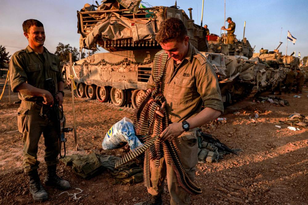 An Israeli soldier walks with an ammunition belt past battle tanks deployed at a position along the border with the Gaza Strip and southern Israel. Photo by Menahem Kahana/AFP