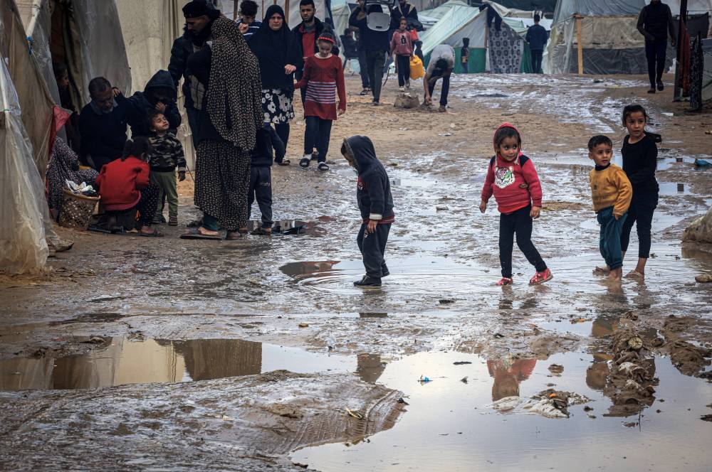 Palestinian children walk in puddles on a muddy path at a makeshift camp housing displaced Palestinians, in Rafah in the southern Gaza Strip, amid Israeli attacks on Gaza on Jan 2, 2024. - Photo by AFP