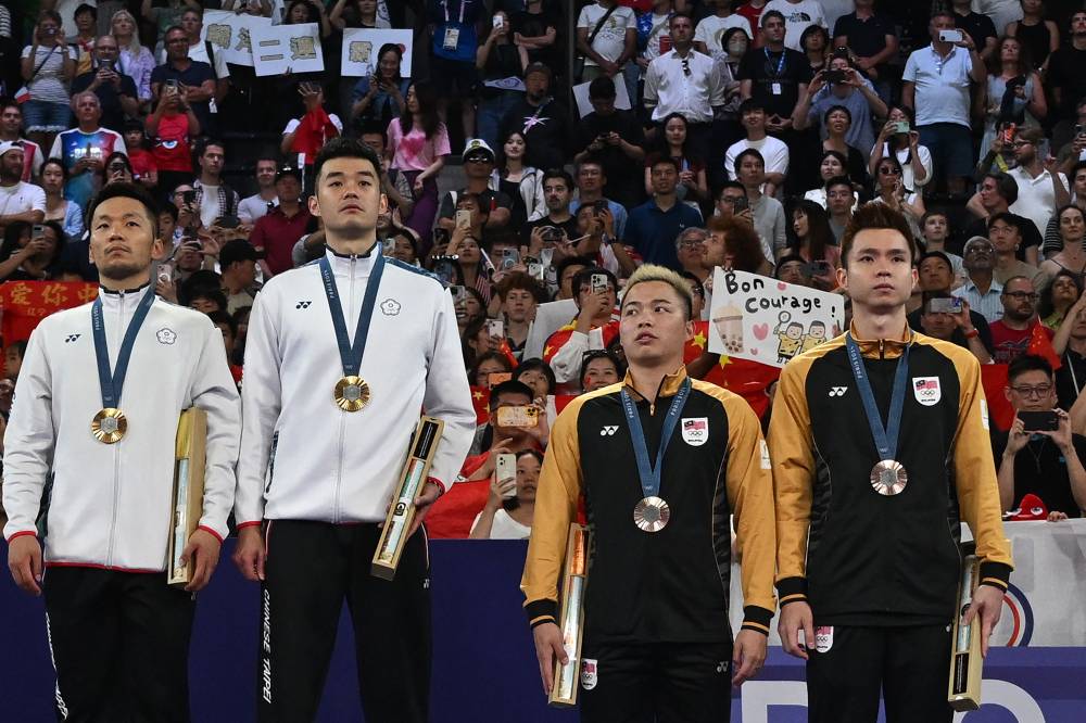 Taiwan's gold medallists (L) Lee Yang and Wang Chi-lin and Malaysia's bronze medallists Aaron Chia and Soh Wooi Yik stand up for the anthem of Chinese Taipei on the podium at the men's doubles badminton medal ceremony during the Paris 2024 Olympic Games at Porte de la Chapelle Arena in Paris on August 4, 2024. (Photo by AFP)