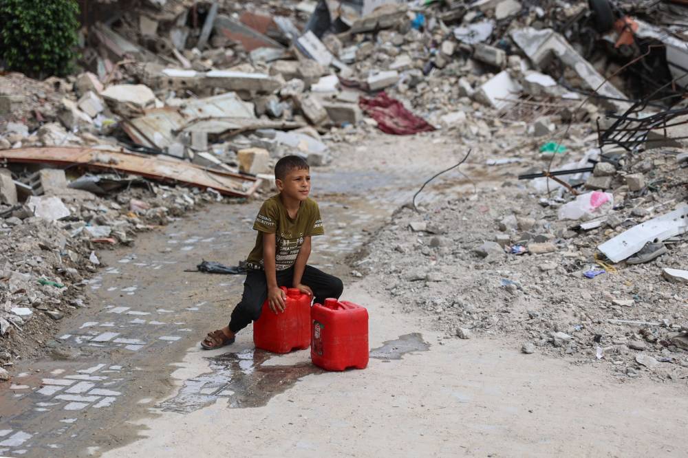 A Palestinian boy rests on water containers, filled from a nearby distribution point, on a dirt road lined with building rubble in the Shujaiya neighbourhood of Gaza City on October 7, 2024, on the first anniversary of the ongoing genocide in the Gaza Strip. (Photo by AFP)