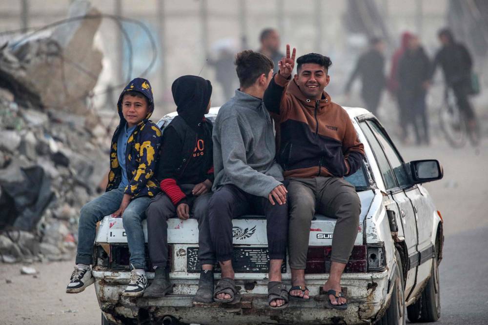 A youth gestures while sitting with other children on the boot of a moving vehicle as people gather along the Salah al-Din road in Nuseirat near the Netzarim corridor waiting to cross to the northern part of the Gaza Strip on Jan 27, 2025. - (Photo by EYAD BABA / AFP)