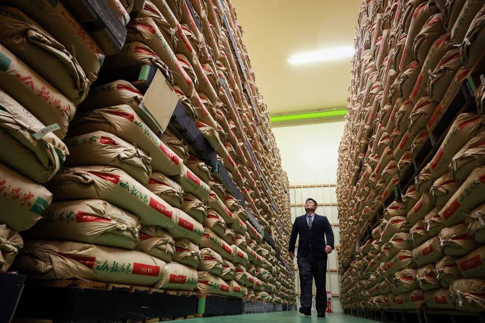 This file photo taken on February 18, 2025 shows a man walking past sacks of government stockpiled rice piled up in a warehouse in Saitama prefecture, north of Tokyo. (Photo by JIJI Press/AFP)