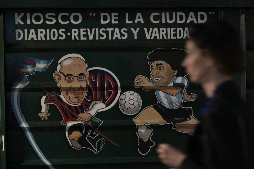 A woman walks past a newsstand with paintings depicting late Pope Francis (L) dressed as a San Lorenzo football club player and late football legend Diego Maradona near the Buenos Aires Cathedral on April 22, 2025. (Photo by JUAN MABROMATA / AFP)