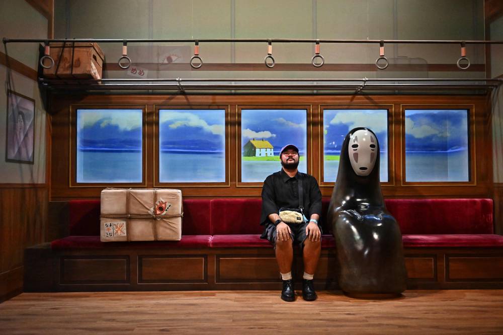This file photo taken on June 29, 2023 shows a man sitting next to the character No Face from the Studio Ghibli film "Spirited Away" during a media preview for "The World of Studio Ghibli's Animation Exhibition Bangkok" before the public opening on July 1, in Central World shopping mall in Bangkok. - (Photo by Lillian SUWANRUMPHA / AFP)