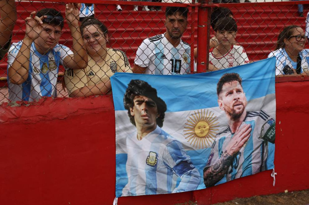 Argentina's national football fans with a flag with the picture of late Argentine football legend Diego Maradona and Forward Lionel Messi attend a training session with the U20 national team at the Tomas Adolfo Duco Stadium in Buenos Aires, on March 22, 2025. - (Photo by ALEJANDRO PAGNI/AFP)