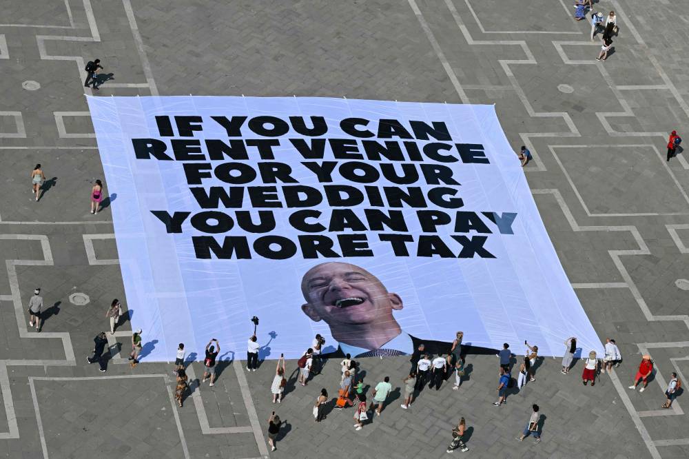 Activists of international environmental group Greenpeace deploy a giant banner displaying a picture of Jeff Bezos and reading "If you can rent Venice for your wedding you can pay more tax" at St Mark square in Venice on June 23, 2025. (Photo by Stefano Rellandini / AFP)