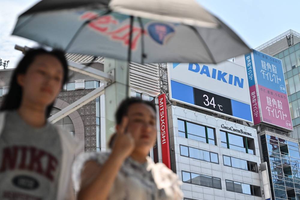 Pedestrians use an umbrella to shelter from the sun as they walk past a digital display (behind) showing a late morning temperature at 34 Centigrade (93.2 F) in the Ginza area of central Tokyo on July 1, 2025. (Photo by Richard A. Brooks / AFP)