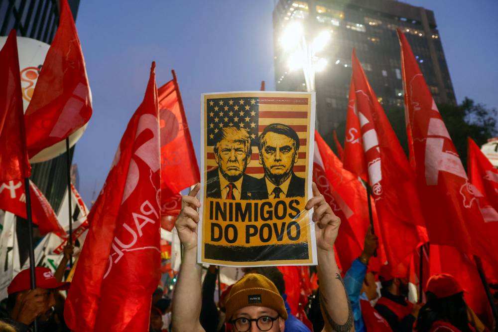 A man holds a sign with images of US President Donald Trump and former Brazilian President Jair Bolsonaro that reads "Enemies of the people" during a demonstration calling to tax the super-rich and demanding the end of the six-day workweek. Photo by Miguel Schincariol/AFP FIL EPIX