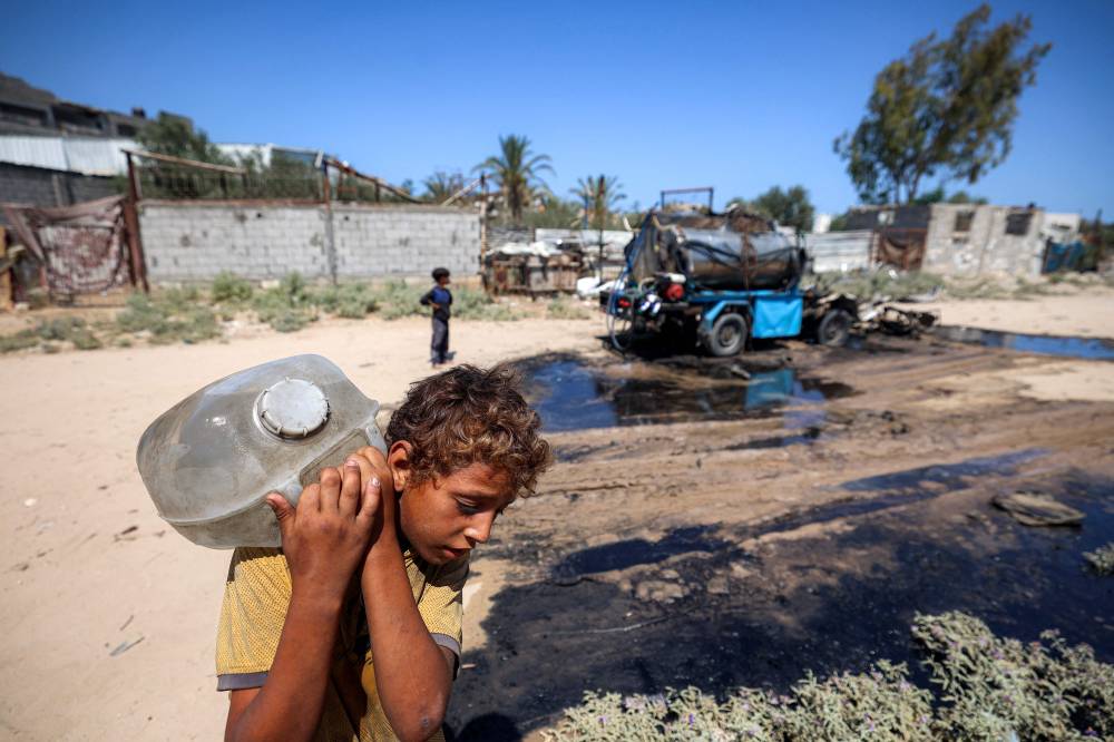 A boy carries on his back a jerrycan filled with water drawn from the tank of a destroyed mobile water cistern that was hit by Israeli bombardment in the Nuseirat camp for Palestinian refugees in the central Gaza Strip on July 14, 2025. (Photo by Eyad BABA / AFP)