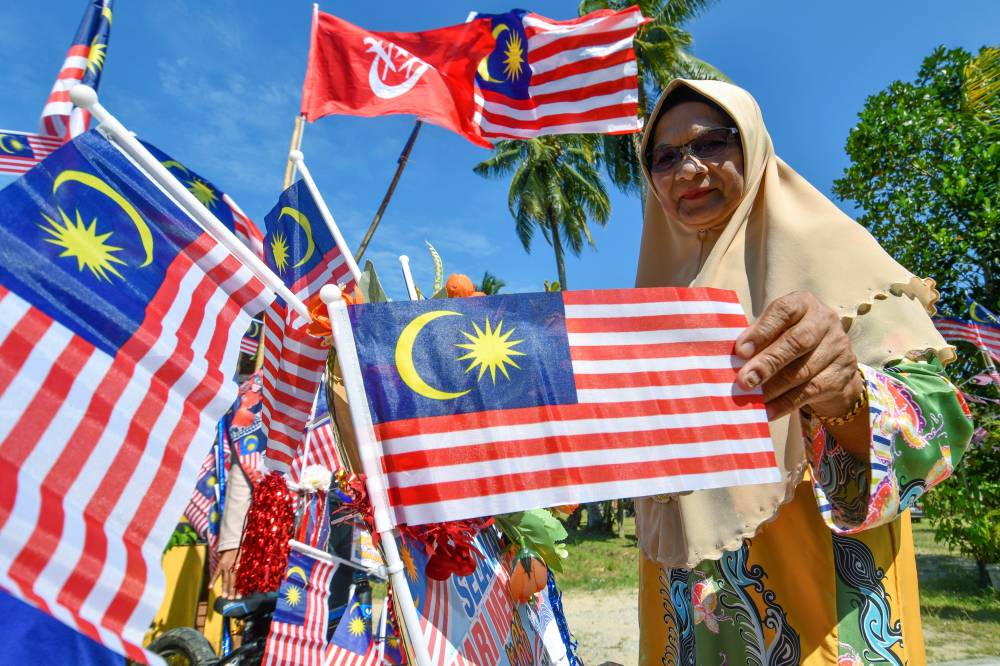 Tuan Yah waving a Jalur Gemilang among the flags that adorn the area around her home in Kampung Kubang Bongor, Melor. She carries on her late husband’s tradition of flag raising. - Bernama photo