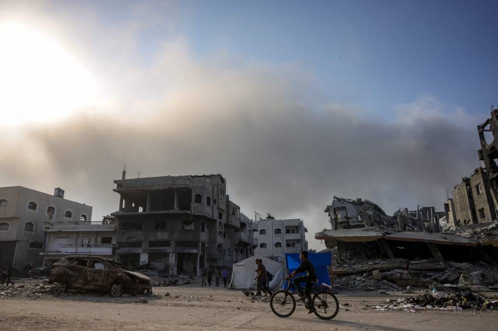 A Palestinian youth cycles past the rubble of destroyed buildings in Jabalia, in the northern Gaza Strip, as smoke billows on the horizon during Israeli bombardment on Aug 25, 2025. - (Photo by BASHAR TALEB / AFP)