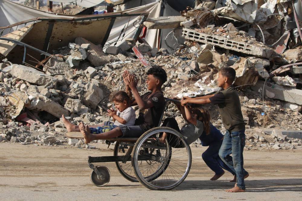 Displaced Palestinian children push two others on a wheelchair past a destroyed building at the Bureij camp for refugees in the central Gaza Strip on Oct 6, 2025. Photo by Eyad Baba/AFP