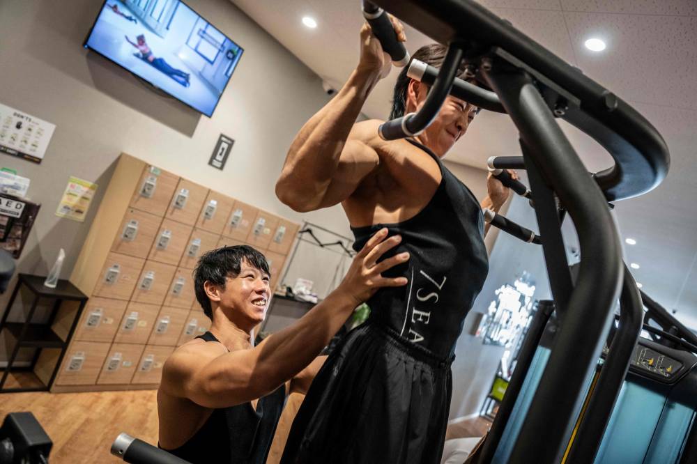 In this photograph taken on September 29, 2025, care workers and bodybuilders Takuya Usui (L) and Hokuto Tatsumi train at a gym in Ichinomiya, Aichi prefecture. Gender stereotypes and poor pay have long discouraged young men from becoming caregivers, even as demand surges in a country with one of the world's oldest populations. But Visionary, a firm based in the city of Nagoya, is successfully recruiting male bodybuilders to the care industry -- luring them to its promised 