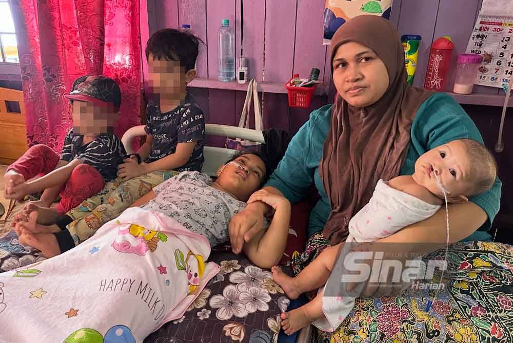 Nik Nur Atiqah (right) cradles her baby, Nur Eleena Huwaina, while holding the hand of her bedridden daughter, Nur Elyana Umairah, as her two other children sit beside them. - PHOTO: Sinar / Adila Sharinni Wahid
