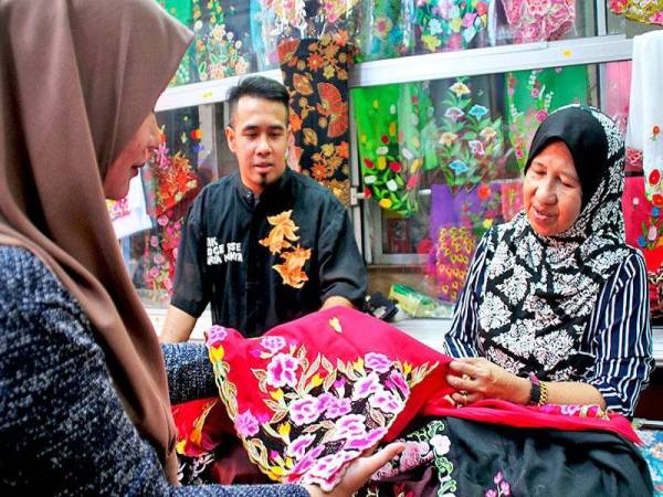 EMMY (kanan) dan Ezrizikri (tengah) menerangkan corak sulaman kebaya nyonya kepada pengunjung Butik Toko Ce Rose, Bandar Hilir, Melaka.