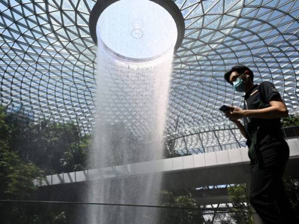 Seorang lelaki memakai topeng muka berjalan melepasi paparan Rain Vortex di Lapangan Terbang Changi. Foto: AFP