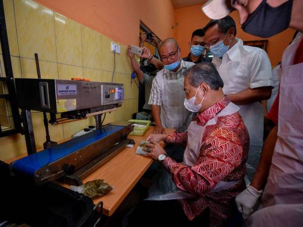 Muhyiddin (duduk) cuba melakukan proses membungkus perencah masakan semasa mengadakan lawatan ke De Muara Enterprise di Bukit Gambir hari ini. - Foto Bernama