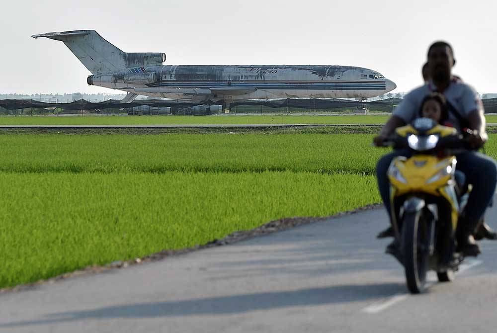 Penduduk setempat menunggang motosikal sambil menikmati keindahan pemandangan sawah padi berlatar belakangkan sebuah kapal terbang yang tidak digunakan lagi. 