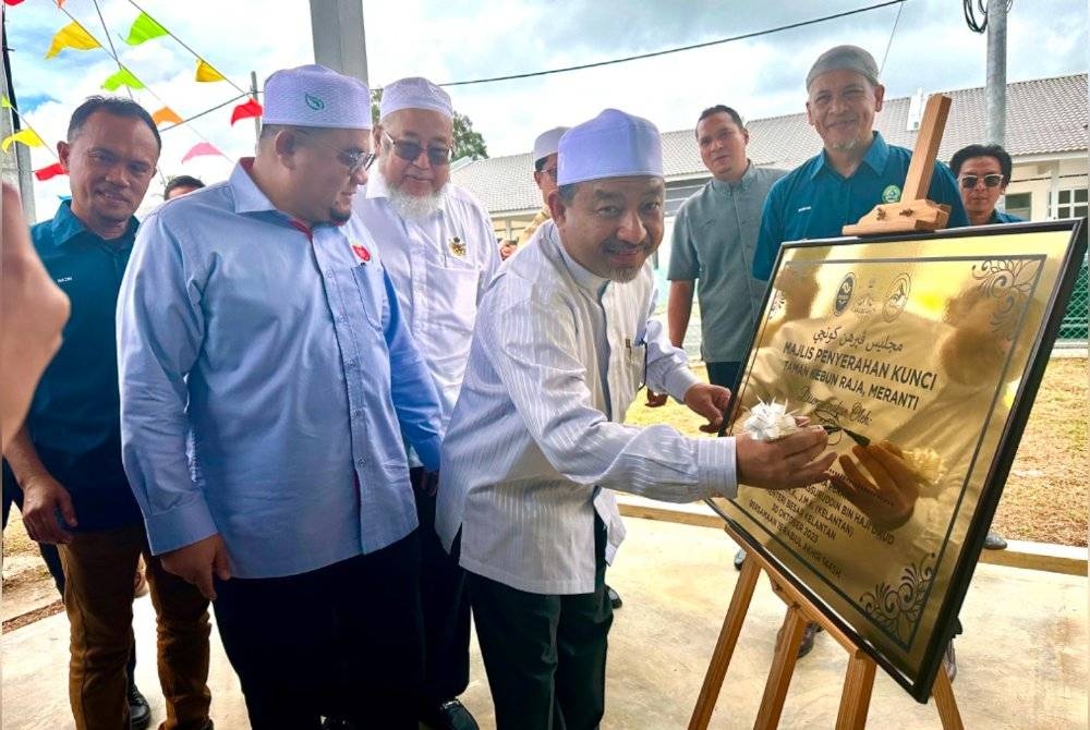 Mohd Nassuruddin (tengah) menurunkan tandatangan ke atas plak bagi simbolik perasmian dan penyerahan kunci RMMK di Meranti, Pasir Mas pada Isnin.