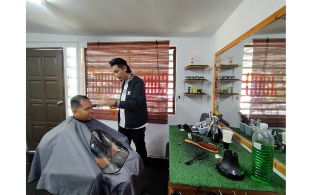 Muhammad Irsyaduddin memotong rambut seorang pelanggannya di kedainya di Kampung Ulu Chepor, Ipoh. 