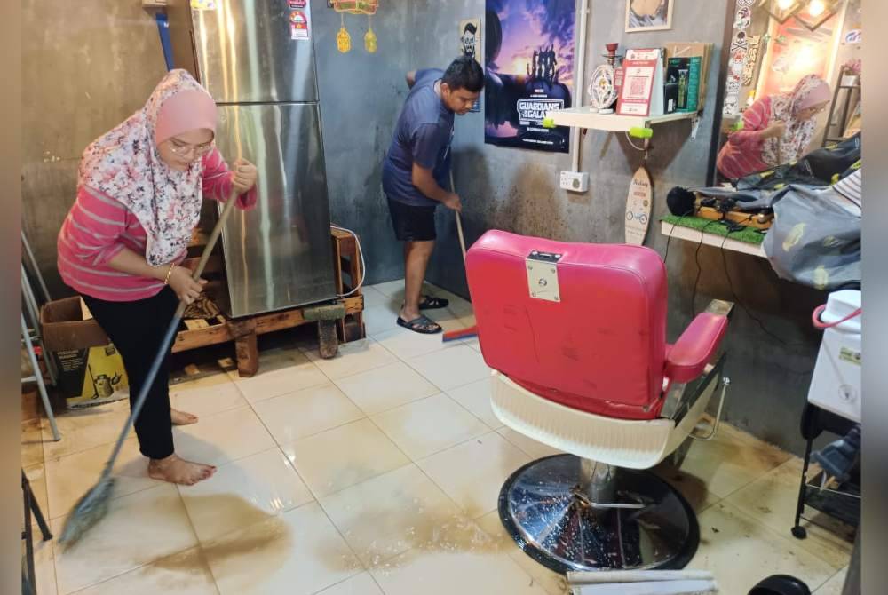 Anis Syazwani (kiri) membantu suaminya membersihkan kedai gunting rambut yang dinaiki banjir kilat.