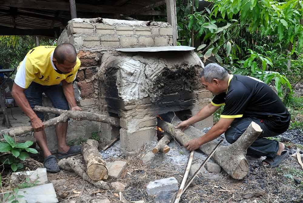 Peniaga lemang, Mohd Zailani Mohd Nor, 49, (kiri) bersama rakannya sedang memasukkan kayu api ke dalam ruang khas pembakaran di dapur relau ciptaannya untuk memasak lemang yang ditempah pelanggan di rumahnya di Kampung Alor Belanga dekat Kampung Alor Keladi.