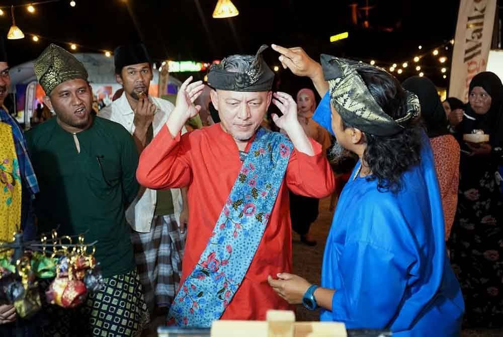 Razali (tengah) mencuba sarung alas kepala atau tanjak sempena Program 'Moreh Selepek Sahur' di Payang Walk di Kuala Terengganu.