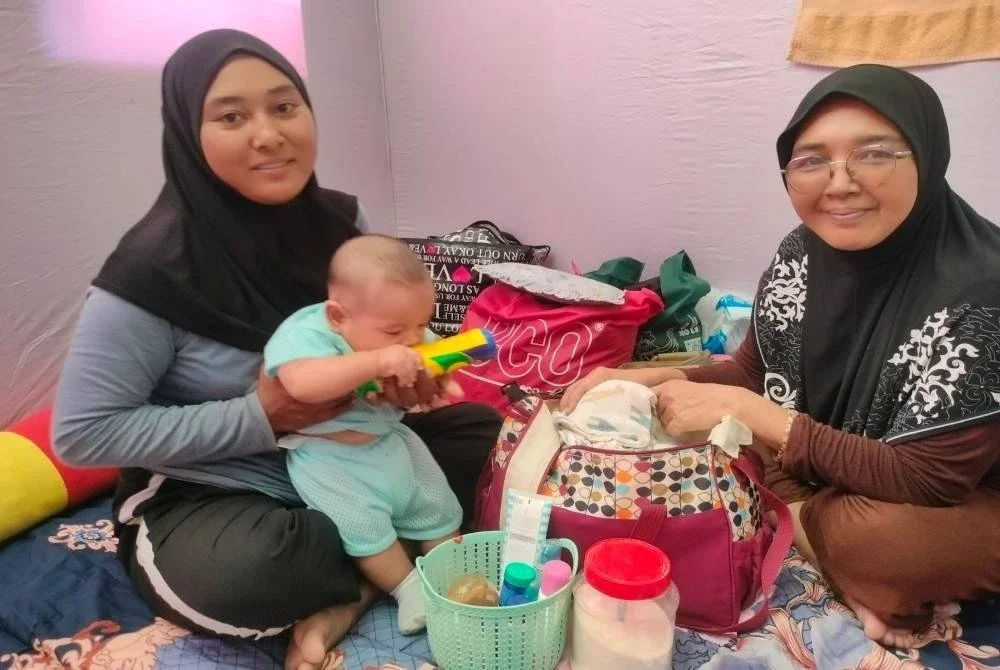 Noratikah (kiri) terpaksa mendukung bayinya yang berusia lima bulan di PPS SK Melayu Raya.