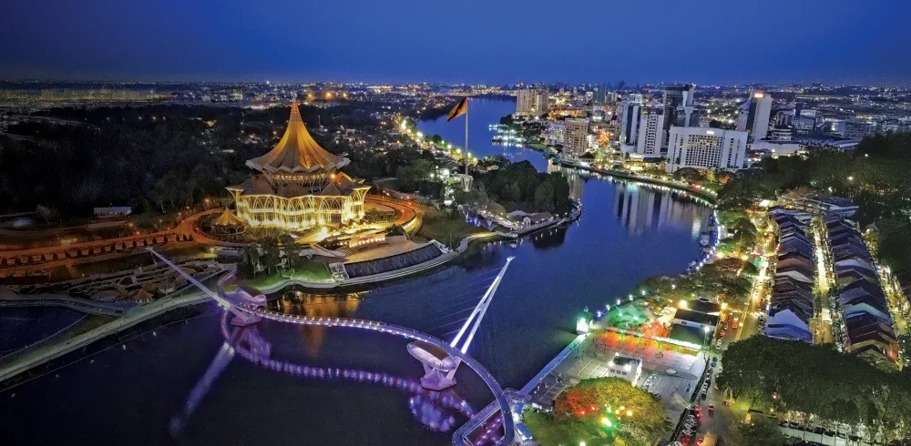 Panorama malam Kuching menampilkan keindahan Dewan Undangan Negeri Sarawak yang bercahaya, terserlah di tepi Sungai Sarawak bersama jambatan ikonik yang memukau.