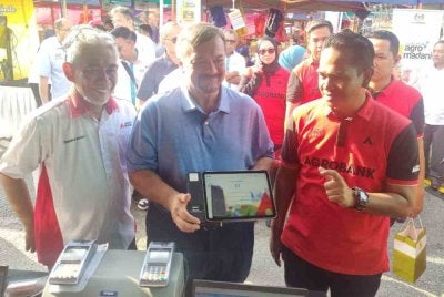 Amir Hamzah (tengah) dan Tengku Ahmad Badli (kiri) menunjukkan peranti Agro Touch iaitu sistem perbankan digital mudah alih semasa lawatan dan ramah mesra bersama penjaja di Pasar Tani Kelana Jaya pada Sabtu.
